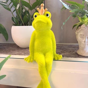 Könnte beinhalten: Eine leuchtend lindgrüne Froschfigur mit einer flauschigen, beflockten Textur. Der Frosch sitzt mit gekreuzten Beinen und trägt eine kleine, glatte, goldfarbene Metallkrone auf dem Kopf. Der Frosch hat große, schwarze Augen.