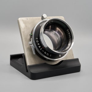 May include: A silver and black camera lens with the text "Copal No. 1" and "Schneider Kreuznach" printed on the side. The lens is mounted on a black stand.