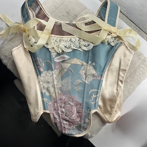 以下が含まれることがあります： 花柄刺繍が施された、ティールとベージュのコルセットトップ。 スウィートハートネックライン、繊細なレーストリム、クリーム色のリボンストラップが特徴で、リボン結びになっています。 コルセットはサテンと柄物の生地の組み合わせです。