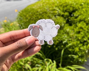May include: A sticker featuring a cartoon sheep with a white body, brown face, and a pink bow. The sticker has the text "I left the 99 to rescue me". The sticker is held in a hand against a background of green foliage.