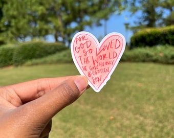 Puede incluir: Una pegatina en forma de corazón rosa con el texto "Porque Dios amó tanto al mundo, que dio a su Hijo unigénito." La pegatina se sostiene en una mano contra un fondo borroso de hierba verde y árboles.