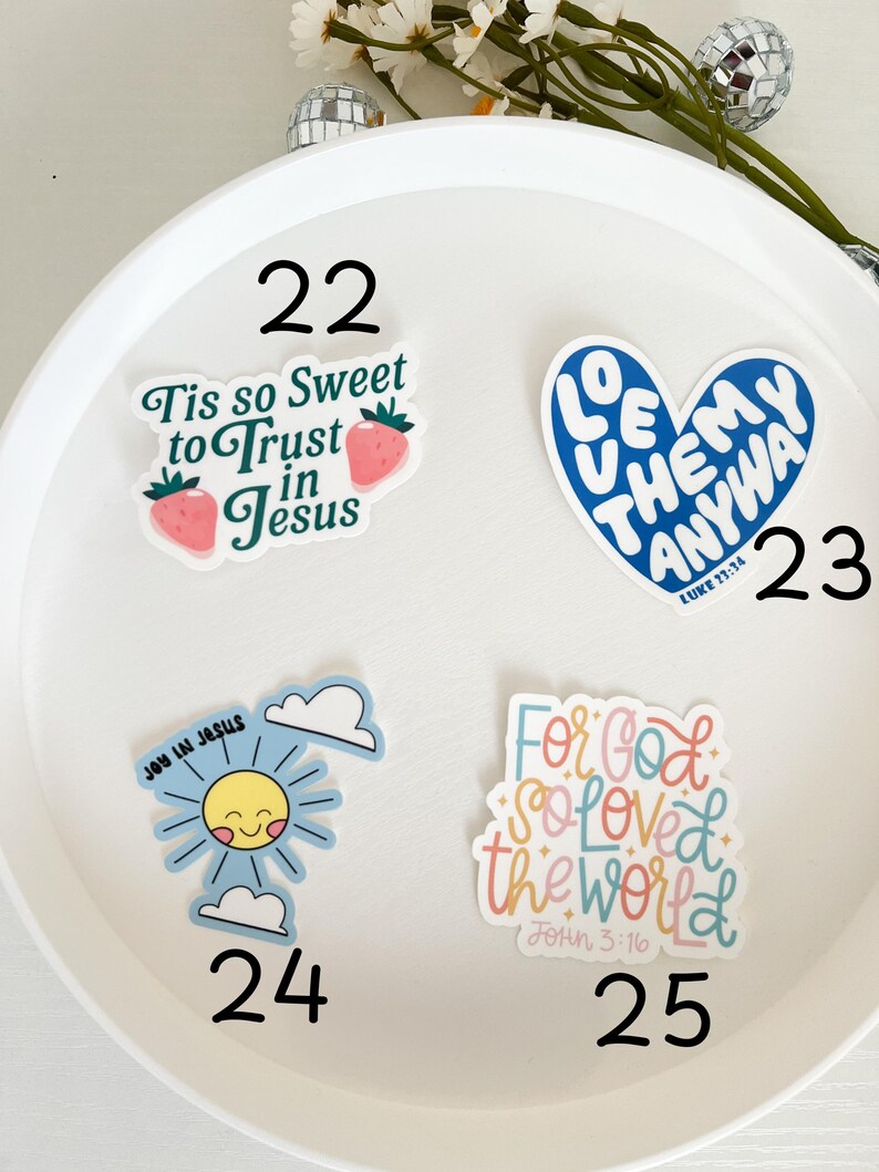 May include: A white plate with four colorful stickers with religious quotes. The stickers are numbered 22, 23, 24, and 25. The stickers feature a strawberry, a heart, a sun, and a hand-drawn font.