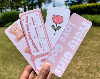 Puede incluir: Tres marcadores de libros rosas y blancos con diferentes diseños. El primer marcador presenta un osito de peluche con un lazo rosa. El segundo marcador tiene una flor rosa con un tallo verde. El tercer marcador tiene el texto "Pretty Girls" en letras blancas sobre un fondo rosa.