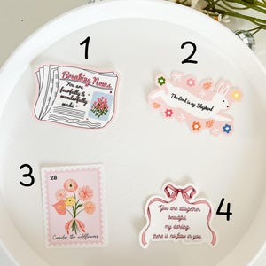 May include: Four pink and white stickers with inspirational quotes and illustrations. The first sticker says "Breaking News: You are fearfully and wonderfully made." The second sticker has a bunny with the quote "The Lord is my Shepherd." The third sticker has a bouquet of flowers with the quote "Consider the wildflowers." The fourth sticker has a pink ribbon with the quote "You are altogether beautiful my darling: there is no flaw in you."