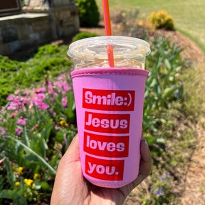 May include: Pink insulated cup holder with the text "Smile :) Jesus loves you." in red letters.