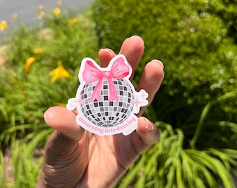 May include: A white sticker with a pink bow on top of a silver disco ball. The sticker says "mourning into dancing".