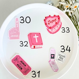 May include: A white plate with pink and red stickers on it. The stickers feature religious themes, including a cross, a heart with the word "Jesus" on it, and a ticket that says "Jesus Fan Club". The plate also has the numbers 30, 31, 32, 33, and 34 written on it.