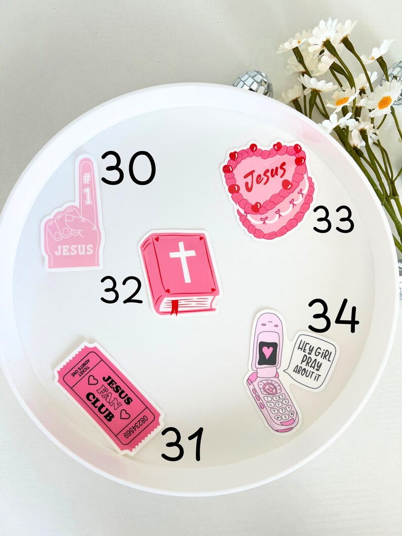 May include: A white plate with pink and red stickers on it. The stickers feature religious themes, including a cross, a heart with the word "Jesus" on it, and a ticket that says "Jesus Fan Club". The plate also has the numbers 30, 31, 32, 33, and 34 written on it.