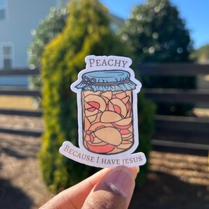 May include: A peach-colored sticker with a cartoon illustration of a jar filled with peaches. The jar has a blue lid and a brown ribbon. The text "Peachy" is above the jar and "Because I have Jesus" is below the jar.