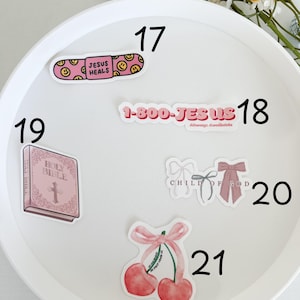 May include: A white plate with pink and white stickers with religious themes. The stickers include a band-aid with the text "Jesus Heals", a phone number with the text "1-800-JESUS", a pink bible with a cross, a pair of pink bows with the text "Child of God", and a pair of cherries with a pink bow and the text "We just love you".
