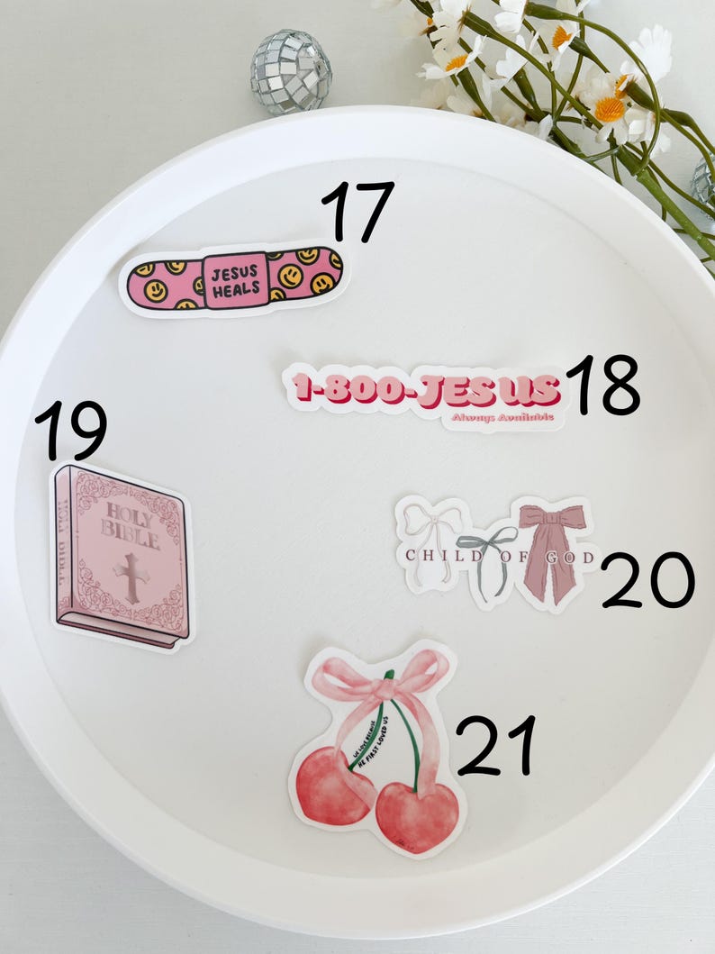 May include: A white plate with pink and white stickers with religious themes. The stickers include a band-aid with the text "Jesus Heals", a phone number with the text "1-800-JESUS", a pink bible with a cross, a pair of pink bows with the text "Child of God", and a pair of cherries with a pink bow and the text "We just love you".