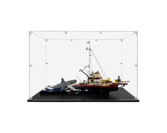 May include: A detailed model of a boat and a shark, displayed inside a clear acrylic case. The boat is primarily red, white, and brown, with a tall mast and various details. The shark is gray and appears to be attacking the boat.