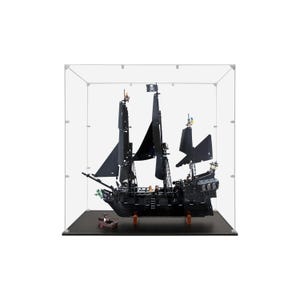 May include: A black pirate ship model with black sails and a skull and crossbones flag, displayed inside a clear acrylic case. The model includes a small boat and is set on a black base.