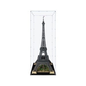 May include: A detailed model of the Eiffel Tower, constructed from interlocking blocks, displayed within a clear protective case. The gray tower features intricate lattice work and is set on a base with miniature trees and structures. The top of the tower has a small French flag.