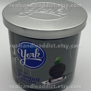 Puede incluir: Una vela de vidrio en un frasco con tapa plateada y una etiqueta azul y blanca. La etiqueta dice "York" en un círculo azul y "Vela perfumada de triple mecha" con una imagen de un caramelo York Peppermint Patty. El frasco es de 14 onzas.