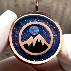 May include: Round pendant with a copper-colored frame. The pendant features a mountain silhouette within a circle, set against a dark blue, starry background. A colorful, iridescent stone is set above the mountain. The pendant has a copper-colored loop.