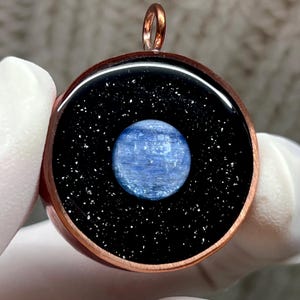 May include: A round pendant featuring a central circular blue kyanite stone set against a black background with silver glitter flecks, all encased in a polished copper frame with a small circular bail at the top for hanging on a chain.