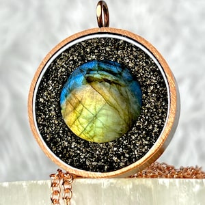 May include: A circular pendant featuring a central round labradorite gemstone with blue and yellow iridescence. The stone is surrounded by a textured black granular border, encased in a copper-toned metal frame with a small top bail.