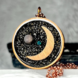 May include: A gold crescent moon pendant necklace with a black and gold glitter background. The pendant features a small, white opal and a green stone.