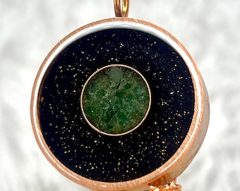 May include: A circular pendant featuring a central green stone inlay surrounded by a black field with gold-colored flecks. The piece is encased in a brushed copper-toned metal frame with a matching metal bail at the top and a link chain attached.