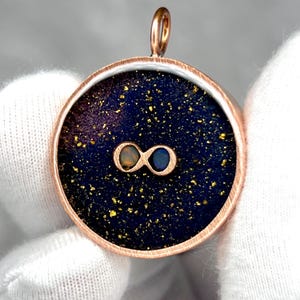 May include: A round pendant with a copper-colored frame. The pendant features a dark blue background with gold flecks and two infinity symbols. The infinity symbols are in shades of orange and blue. The pendant has a loop at the top for a chain.