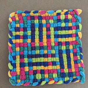 May include: A colorful woven pot holder with a square shape and a scalloped edge. The pot holder is made with a variety of colors, including blue, green, yellow, pink, and purple.
