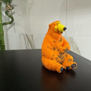 May include: A small, seated bear figurine with a bright orange body, a yellow snout, and dark blue accents on the nose, eyes, and paw pads. The bear has a textured surface with dark brown patches across its chest and legs, and it is posed in a sitting position with its front paws resting on its belly.