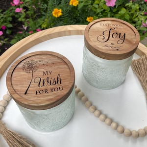 May include: Two glass jars with textured surfaces and round wooden lids. One lid features a dandelion graphic and the text "MY WISH FOR YOU". The other lid features a floral graphic and the text "FIND YOUR JOY". The jars are placed on a white surface.