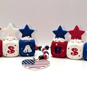 May include: A patriotic display featuring red, white, and blue decorative items. The centerpiece is a series of cube-shaped ornaments spelling "USA", topped with star-shaped accents. A heart-shaped decoration with a flag design and a beaded garland complete the arrangement.