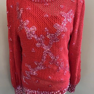 Peut inclure: Un pull rouge en maille ajourée à manches longues. Le pull est orné de paillettes roses formant un motif floral sur le devant et autour de l'encolure et des poignets. Le pull est présenté sur un mannequin.