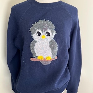 Pode incluir: Camisola azul marinho com um desenho de coruja bordada. A coruja tem penas cinzentas e brancas, olhos pretos grandes e um bico e pés amarelos. A coruja está empoleirada num galho castanho.