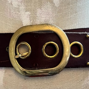 Puede incluir: Un cinturón de cuero marrón oscuro con una hebilla ovalada dorada grande. El cinturón presenta ojales y remaches dorados. Este cinturón es un accesorio elegante.