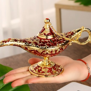 Peut inclure: Une lampe à genie dorée et rouge avec des détails complexes et une chaîne suspendue au sommet. La lampe est posée sur la main d'une personne.
