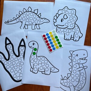 Op de afbeelding: Vijf zwart-witte dinosaurus contouren met cirkels voor stippen stickers. De dinosaurussen omvatten een stegosaurus, een triceratops, een brachiosaurus, een voetafdruk en een tyrannosaurus rex. Een vel met gekleurde stippen stickers is inbegrepen.