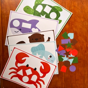 May include: A set of five printable activity cards featuring various sea creatures, including a turtle, a dolphin, an otter, a fish, and a crab. Each card has a shape cutout for children to match with the corresponding shape from a separate set of colourful shapes.