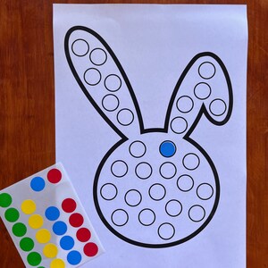K&ouml;nnte beinhalten: Ein druckbares Punkt-zu-Punkt-Aktivit&auml;tsblatt in Form eines Hasen mit einem blauen Punkt in der Mitte des Hasenkopfes. Ein Blatt mit bunten Punkt-Aufklebern ist enthalten.