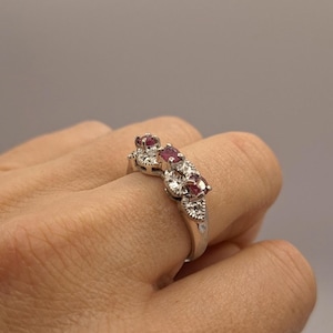 Könnte beinhalten: Ein silberfarbener Ring mit drei oval geschliffenen, dunkelrosa Rubinen in einer Reihe, die jeweils durch kleine Gruppen von klaren, funkelnden, rund geschliffenen Steinen im Metallband getrennt sind und ein durchgehendes Design bilden.