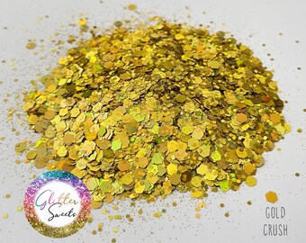 May include: A pile of gold-colored glitter with a mix of hexagonal and irregular shapes. The glitter is scattered on a white surface. The text "Gold Crush" is visible in the image, along with a logo that says "Glitter Sweets".