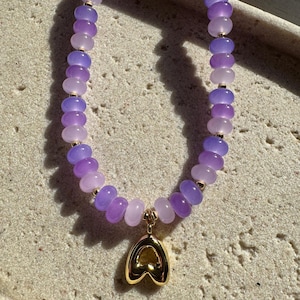 May include: A necklace with lavender and purple beads, interspersed with gold-coloured accents. The necklace has a gold heart-shaped pendant. The beads are smooth and rounded, creating a delicate and elegant appearance.