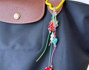 May include: A black handbag with a brown leather flap and a decorative charm. The charm features a yellow loop, colorful strings, and various beads, including a red mushroom, clear beads, and a blue heart.