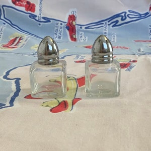 May include: Two clear glass salt and pepper shakers with silver tops. The shakers are sitting on a white tablecloth with a blue and white map design.