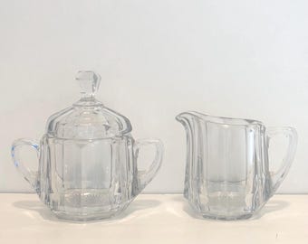 Peut inclure: Un sucrier en verre transparent avec couvercle et une crémière. Les deux pièces présentent un design à facettes et de petites poignées. Le sucrier a un couvercle avec une petite poignée décorative. La crémière a un bec verseur.