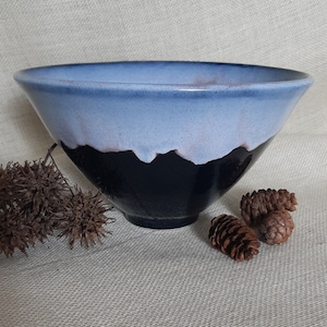 May include: A large, blue and black ceramic bowl with a white rim. The bowl has a unique glaze that creates a mountain-like pattern. The bowl is sitting on a light brown fabric surface.
