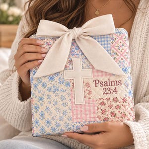 May include: A square quilted fabric book cover featuring a patchwork design of blue and pink floral and gingham patterns. A large cream-colored fabric bow is attached to the top. A white fabric cross and the embroidered text Psalms 23:4 appear.