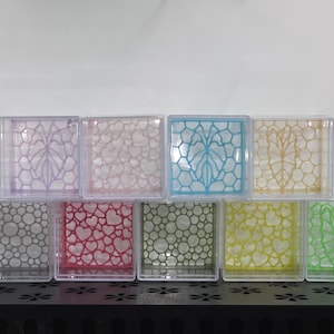 May include: A collection of nine square, clear plastic display boxes arranged in two rows. Each box features a unique, colorful, laser-cut insert with various patterns, including hearts, honeycomb, and leaf-like designs in shades of pink, blue, yellow, green, and gray.