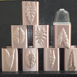 May include: A collection of eight rose gold rectangular containers arranged in a pyramid shape. Each container features a unique embossed leaf design on the front surface. One container on the top right includes a clear plastic dome-shaped lid.