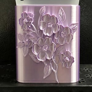 May include: A light purple rectangular container featuring a raised, embossed floral design on the front surface. The flowers and leaves are detailed with a glossy finish. The letter B is printed in the upper left corner, and a translucent white lid sits on top.