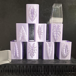 May include: A collection of seven light purple rectangular planters arranged in a pyramid, each featuring a unique embossed leaf design on the front. One clear plastic insert is visible in the top right planter, and a small square base sits nearby.