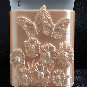 May include: A peach-colored rectangular container featuring a raised relief design of two butterflies and seven daisies on the front surface. The letter D is printed in black in the upper left corner against a translucent top border.