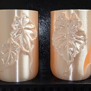 May include: Two cylindrical peach-colored containers featuring embossed leaf designs on their exterior surfaces. The left container displays two overlapping leaves, while the right container features a single large leaf and a vertical cutout detail.